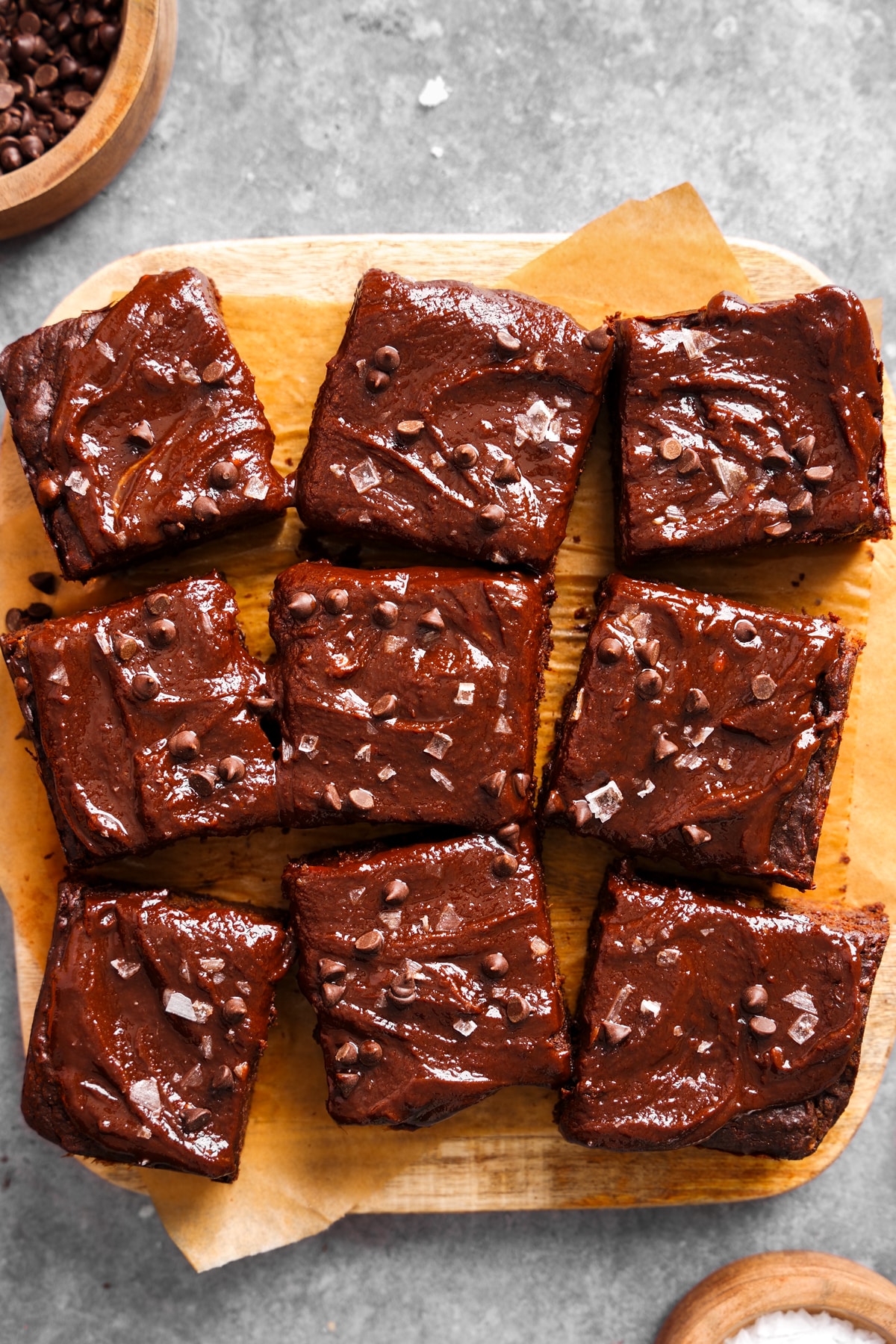 A wooden board topped with nine square vegan chocolate brownies, each covered in glossy chocolate frosting and sprinkled with flaky sea salt and mini chocolate chips. The brownies rest on a piece of parchment paper, with a bowl of chocolate chips visible in the background.