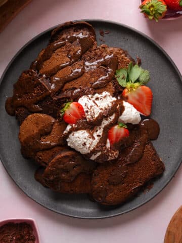 Vegan chocolate brownie pancakes topped with coconut whipped cream, chocolate drizzle, and fresh strawberries on a plate, an easy dairy-free plant-based breakfast recipe.