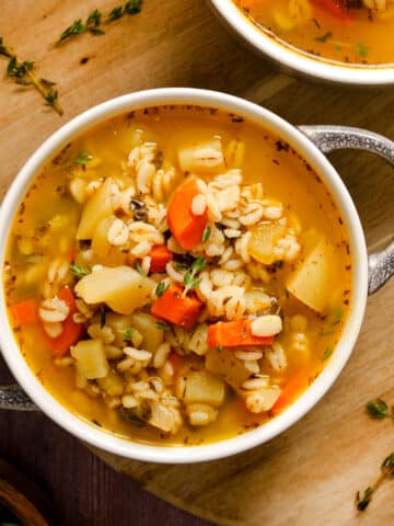 Bowl of hearty vegan barley soup filled with carrots, potatoes, celery, and fresh herbs in a golden vegetable broth, served on a wooden board for a cozy, comforting meal.