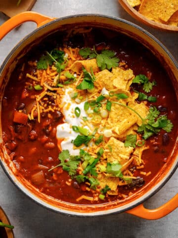 Top-down view of a hearty vegan bean chili in an orange Dutch oven, topped with vegan sour cream, shredded vegan cheese, tortilla chips, green onions, and fresh cilantro on a gray surface.
