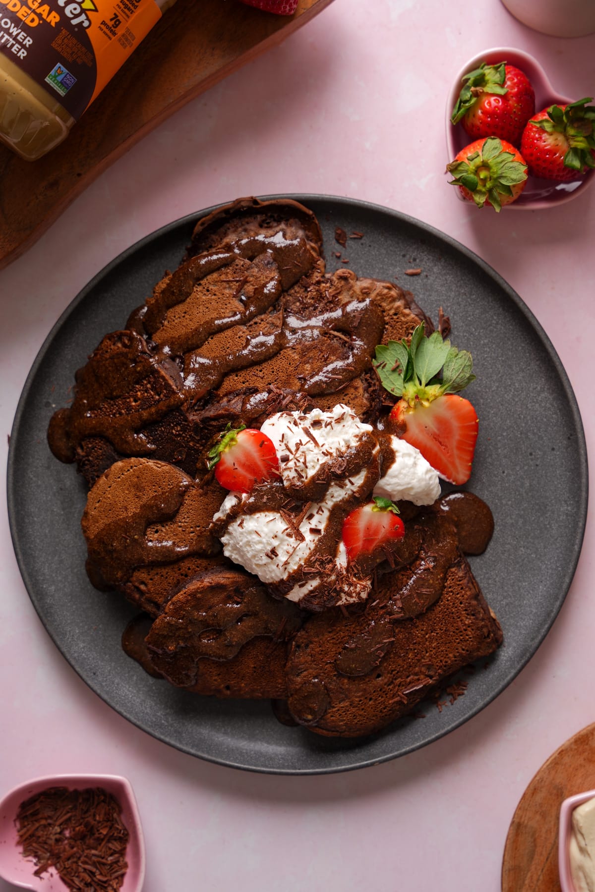 Vegan chocolate brownie pancakes topped with whipped cream, chocolate drizzle, shaved chocolate, and fresh strawberries on a plate, served as a decadent dairy-free plant-based breakfast.