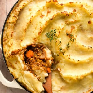 Close-up of vegan lentil shepherd's pie with creamy mashed potato topping, a spoonful scooped out to reveal a hearty lentil and vegetable filling, garnished with fresh thyme in a white baking dish.