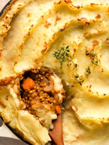 Close-up of vegan lentil shepherd’s pie with creamy mashed potato topping, a spoonful scooped out to reveal a hearty lentil and vegetable filling, garnished with fresh thyme in a white baking dish.