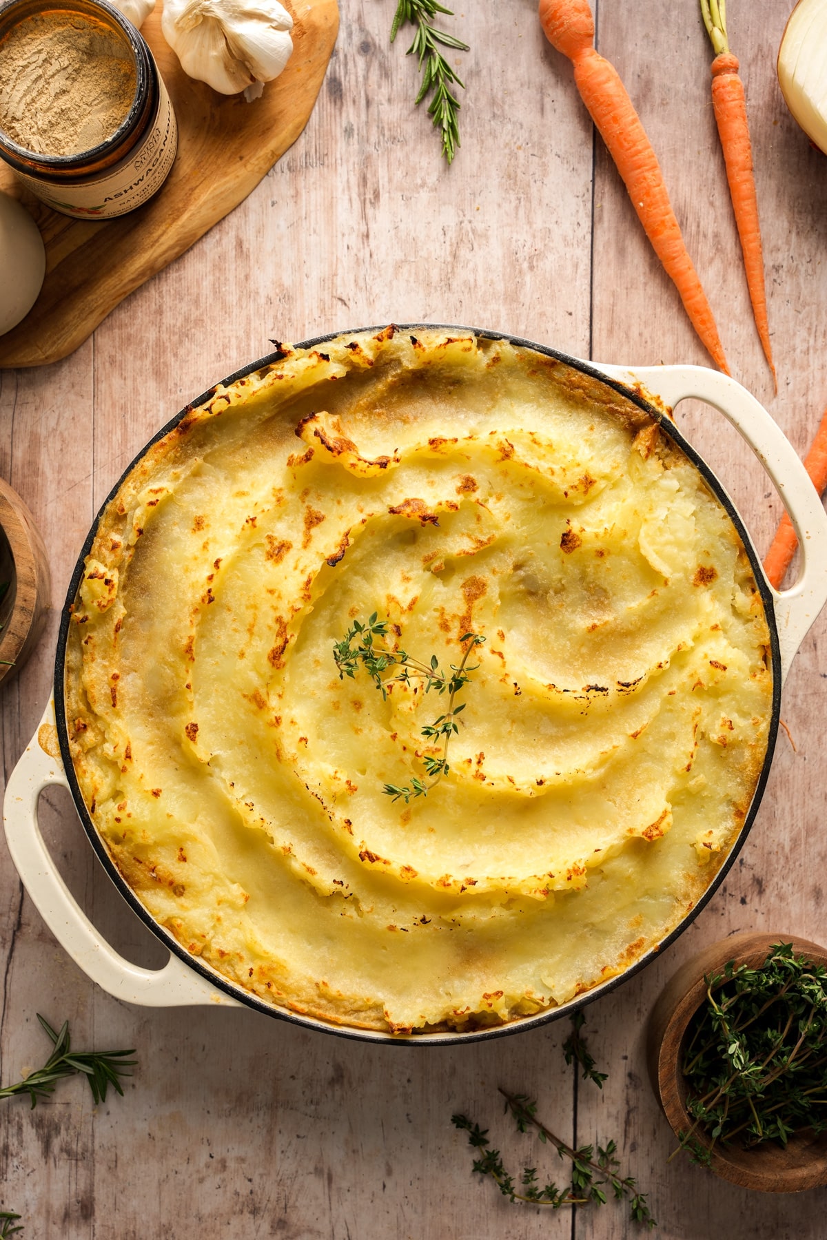 Vegan lentil shepherd's pie topped with golden mashed potatoes, baked in a white casserole dish and garnished with fresh thyme, surrounded by carrots, garlic, and herbs on a rustic wooden surface.