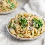 Close-up view of a bowl of creamy vegan cauliflower Alfredo fettuccine topped with broccoli florets, mushrooms, peas, and cracked black pepper. The bean-based fettuccine is coated in a rich, dairy-free cauliflower Alfredo sauce, creating a comforting and nutritious plant-based pasta dish. A second bowl of pasta and a white kitchen towel appear softly blurred in the background, highlighting this healthy vegan dinner recipe.