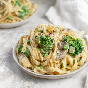 Close-up view of a bowl of creamy vegan cauliflower Alfredo fettuccine topped with broccoli florets, mushrooms, peas, and cracked black pepper. The bean-based fettuccine is coated in a rich, dairy-free cauliflower Alfredo sauce, creating a comforting and nutritious plant-based pasta dish. A second bowl of pasta and a white kitchen towel appear softly blurred in the background, highlighting this healthy vegan dinner recipe.