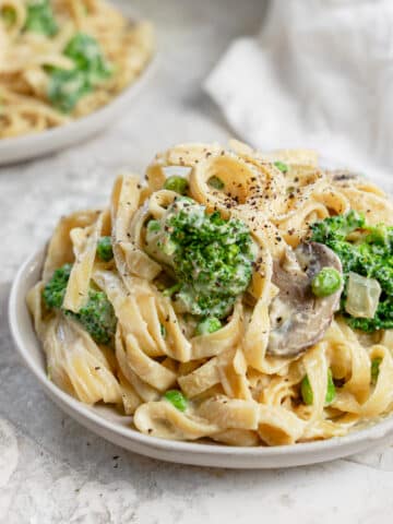 Close-up view of a bowl of creamy vegan cauliflower Alfredo fettuccine topped with broccoli florets, mushrooms, peas, and cracked black pepper. The bean-based fettuccine is coated in a rich, dairy-free cauliflower Alfredo sauce, creating a comforting and nutritious plant-based pasta dish. A second bowl of pasta and a white kitchen towel appear softly blurred in the background, highlighting this healthy vegan dinner recipe.