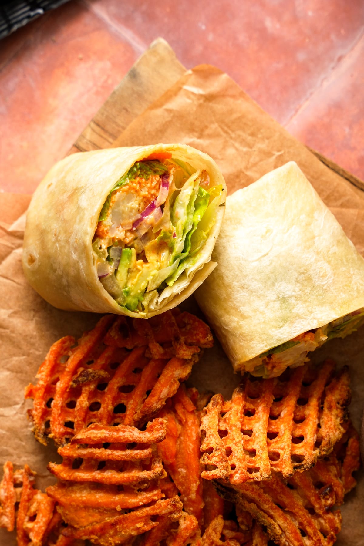 Buffalo cauliflower wrap sliced in half and served with crispy sweet potato waffle fries on parchment paper. The wrap is filled with crispy buffalo cauliflower, fresh romaine lettuce, creamy avocado, red onion, and vegan ranch dressing, creating a colorful and satisfying plant-based meal. The golden tortilla and crispy waffle fries make this healthy vegan lunch or dinner look both comforting and flavorful.