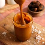 A spoon lifts a thick ribbon of glossy vegan date caramel from a glass jar on a wooden board. Coconut flakes and Medjool dates surround this creamy 2-ingredient, refined sugar-free caramel sauce that's perfect for dipping fruit, drizzling over desserts, or stirring into coffee.