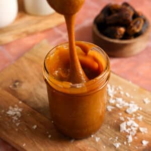 A spoon lifts a thick ribbon of glossy vegan date caramel from a glass jar on a wooden board. Coconut flakes and Medjool dates surround this creamy 2-ingredient, refined sugar-free caramel sauce that's perfect for dipping fruit, drizzling over desserts, or stirring into coffee.