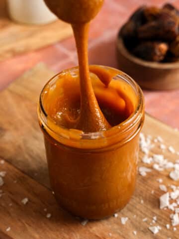 A spoon lifts a thick ribbon of glossy vegan date caramel from a glass jar on a wooden board. Coconut flakes and Medjool dates surround this creamy 2-ingredient, refined sugar-free caramel sauce that's perfect for dipping fruit, drizzling over desserts, or stirring into coffee.