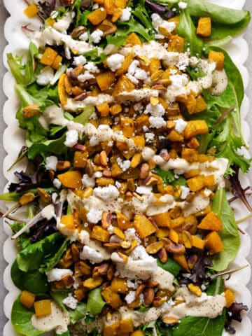 Close-up overhead view of a roasted golden beet salad on a white scalloped serving platter. The salad is made with mixed spring greens and topped with roasted golden beets, crunchy pistachios, crumbled vegan cheese, and a creamy dressing drizzled throughout. The colorful combination of greens, golden beets, and pistachios creates an elegant plant-based salad that's perfect for lunch, dinner, holidays, or entertaining.