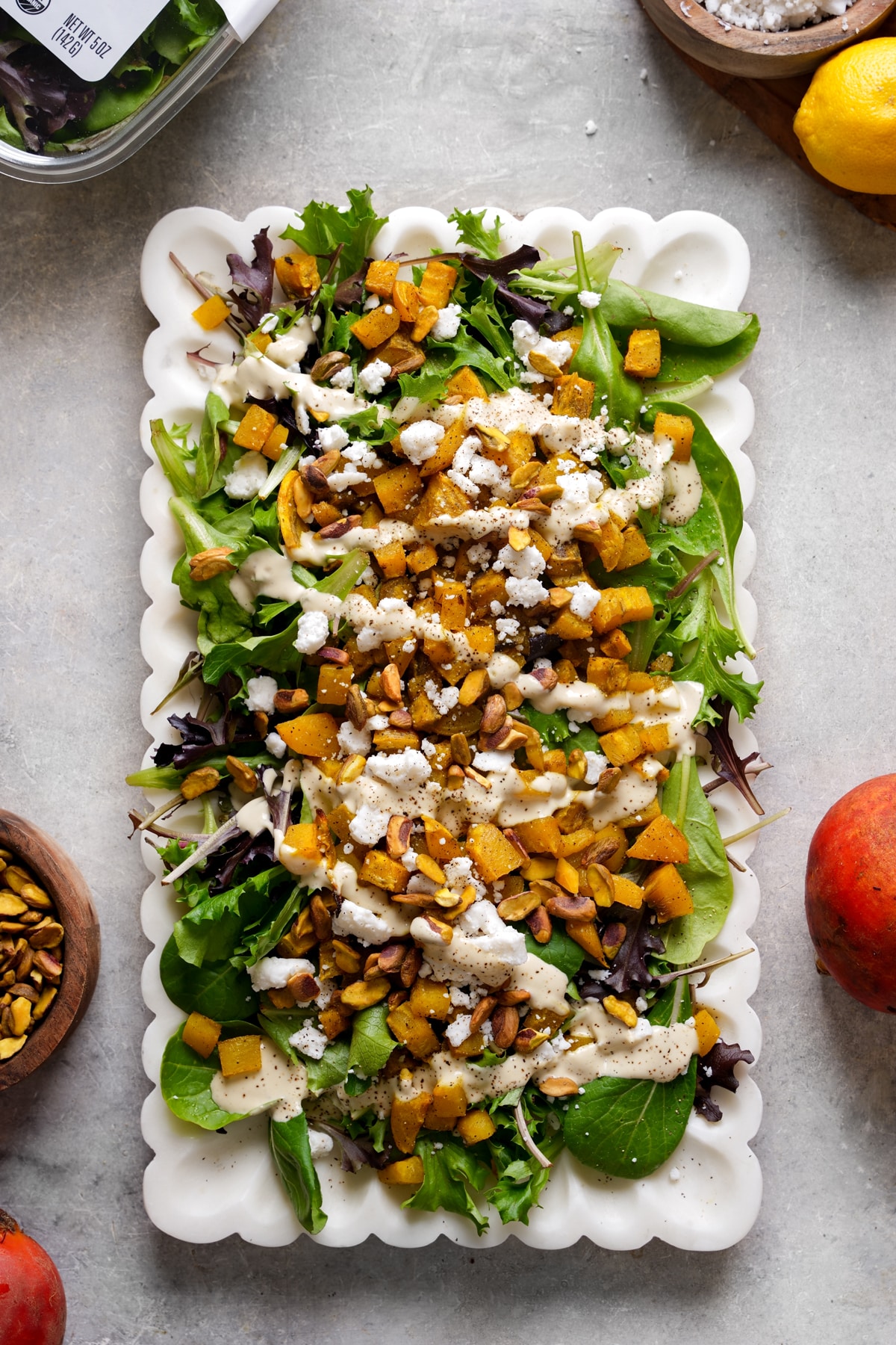Overhead view of a vibrant salad served on a white scalloped platter. The salad features mixed greens topped with roasted golden beets, crumbled vegan cheese, crunchy pistachios, and a creamy dressing drizzled over the top. Fresh peaches, pistachios, a lemon, and additional salad ingredients surround the platter, creating a colorful and elegant plant-based dish perfect for lunch, dinner, or entertaining.