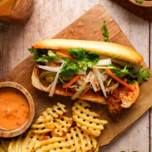 Overhead view of a vegan tempeh bánh mì on a toasted baguette topped with fresh cilantro, pickled carrots and daikon, jalapeños, and spicy mayo, served with crispy waffle fries and a side of creamy dipping sauce on a wooden board.