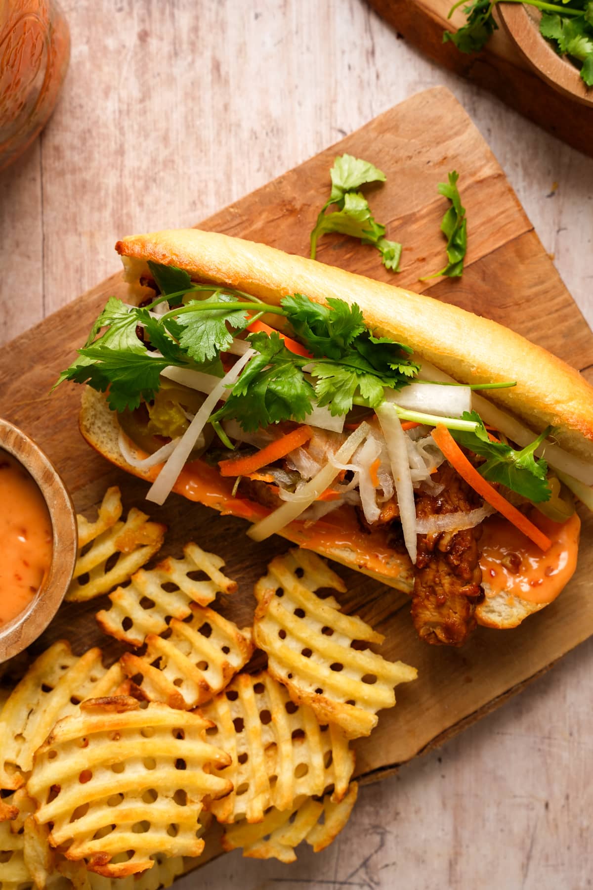Vegan tempeh bánh mì sandwich topped with pickled daikon and carrots, fresh cilantro, jalapeños, and spicy mayo on a toasted baguette, served with crispy waffle fries and dipping sauce on a wooden board.