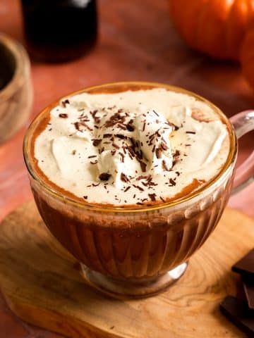 Close-up of vegan pumpkin hot chocolate in a glass mug topped with whipped cream and chocolate shavings, with pumpkins and dark chocolate pieces in the background.