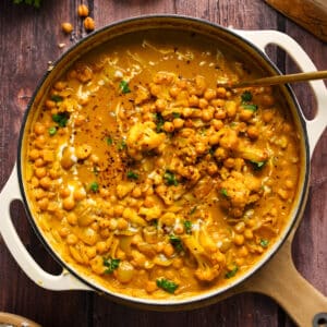 Overhead shot of a creamy vegan coconut chickpea curry simmering in a large cream-colored Dutch oven, filled with chickpeas, cauliflower, onions, and fresh herbs. The golden curry sauce is rich and glossy, swirled with coconut milk and garnished with red pepper flakes and parsley. Wooden bowls of rice, chili flakes, and crispy chickpeas are styled around the pot on a rustic dark wood table, enhancing the cozy, comforting presentation.