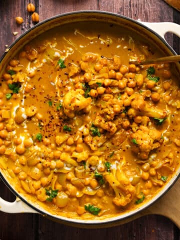 Overhead shot of a creamy vegan coconut chickpea curry simmering in a large cream-colored Dutch oven, filled with chickpeas, cauliflower, onions, and fresh herbs. The golden curry sauce is rich and glossy, swirled with coconut milk and garnished with red pepper flakes and parsley. Wooden bowls of rice, chili flakes, and crispy chickpeas are styled around the pot on a rustic dark wood table, enhancing the cozy, comforting presentation.