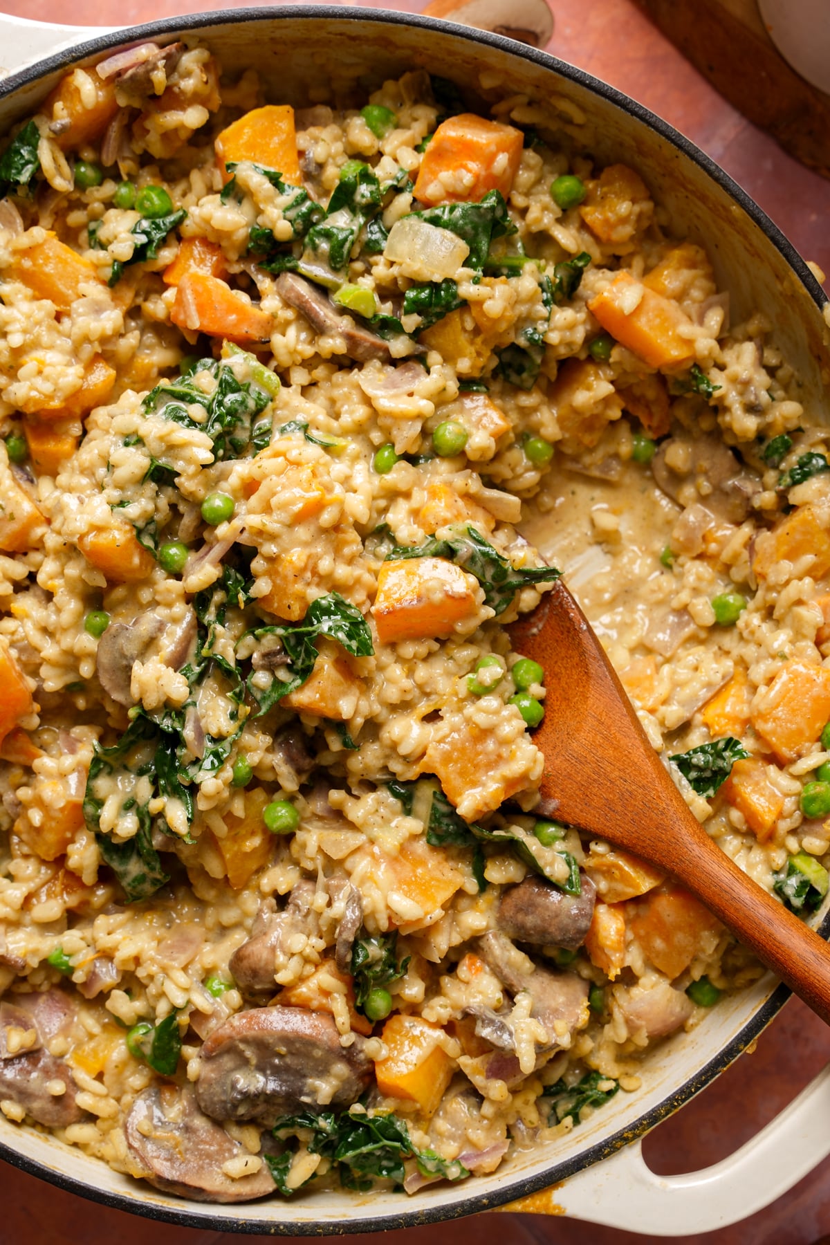 Creamy vegan sweet potato risotto with mushrooms, kale, and green peas in a pot, stirred with a wooden spoon for a cozy one-pot plant-based meal.