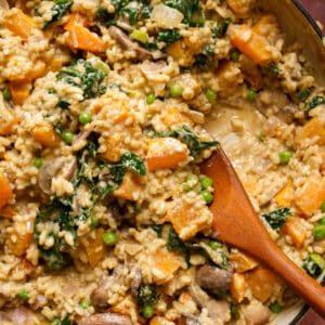 Creamy vegan sweet potato risotto with mushrooms, kale, red onion, and green peas in a pot, served with a wooden spoon.