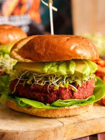 A vibrant vegan beet burger stacked with avocado slices, crisp lettuce, and fresh sprouts on a golden toasted bun, served with a side of sweet potato fries on a rustic wooden board.