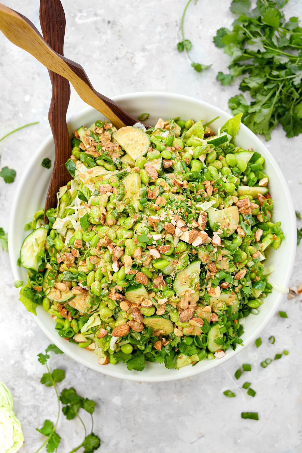 Overhead view of a vibrant vegan edamame crunch salad tossed in creamy peanut miso dressing, served in a large white bowl with sliced cucumbers, cabbage, romaine, cilantro, crushed almonds, and green onions on a light marble background