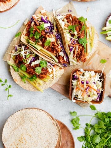 three bbq jackfruit tacos with a bowl of coleslaw on the side and plates with tortillas