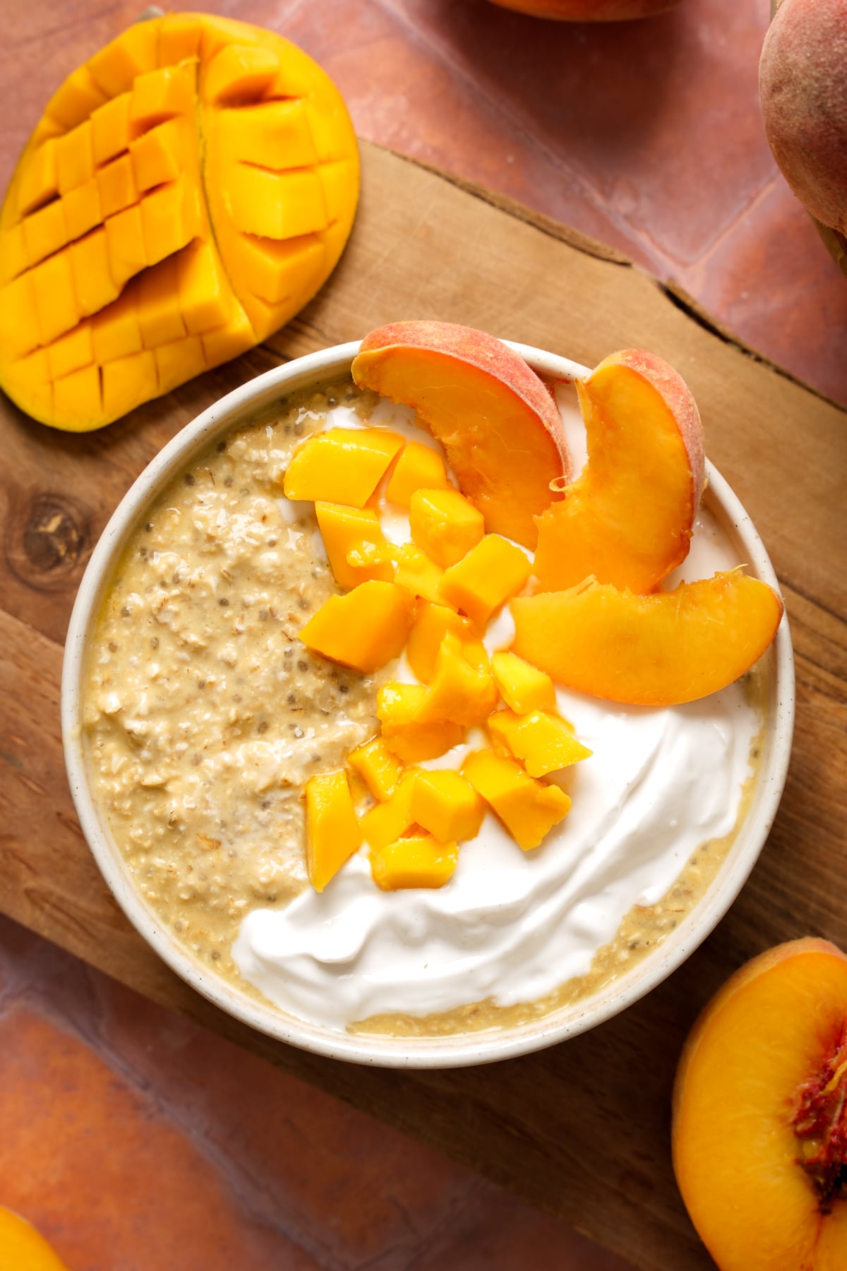A bowl of healthy overnight mango peach oatmeal topped with creamy dairy-free yogurt, fresh diced mango, and sliced peaches. A scored honey mango and whole peaches surround the bowl on a rustic wooden board for a bright, summery breakfast presentation.
