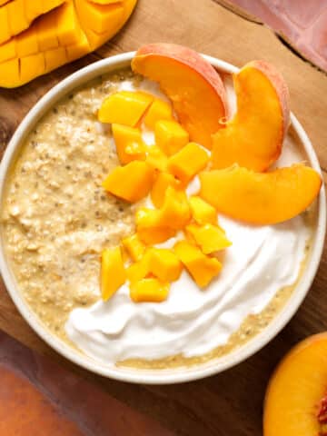 A bowl of healthy overnight mango peach oatmeal topped with creamy dairy-free yogurt, fresh diced mango, and juicy peach slices. Fresh honey mango and ripe peaches surround the bowl on a rustic wooden board, making a bright, easy make-ahead breakfast.
