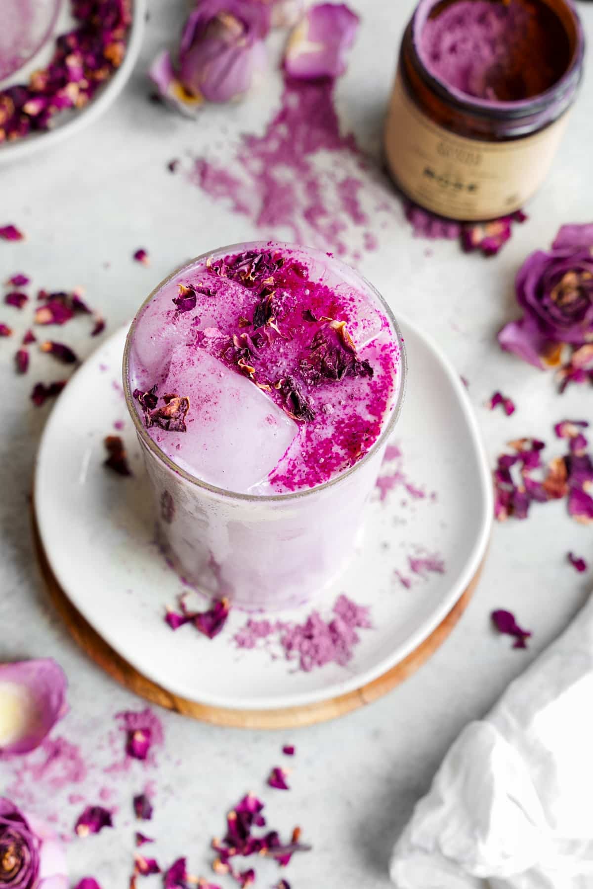 A glass filled with iced vanilla‑rose latte-pale pink and creamy-with vibrant magenta rose powder and dried petals sprinkled on top and around the base on a white plate.