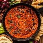 Overhead view of creamy vegan butternut squash dip baked in a cast iron skillet, topped with fresh sage, chives, sesame seeds, and seasoning, surrounded by crackers, bread, grapes, celery, and hummus.