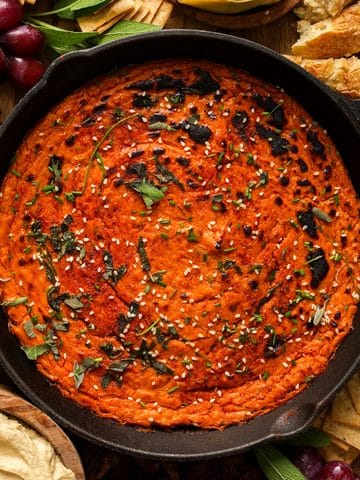 Overhead view of creamy vegan butternut squash dip baked in a cast iron skillet, topped with fresh sage, chives, sesame seeds, and seasoning, surrounded by crackers, bread, grapes, celery, and hummus.