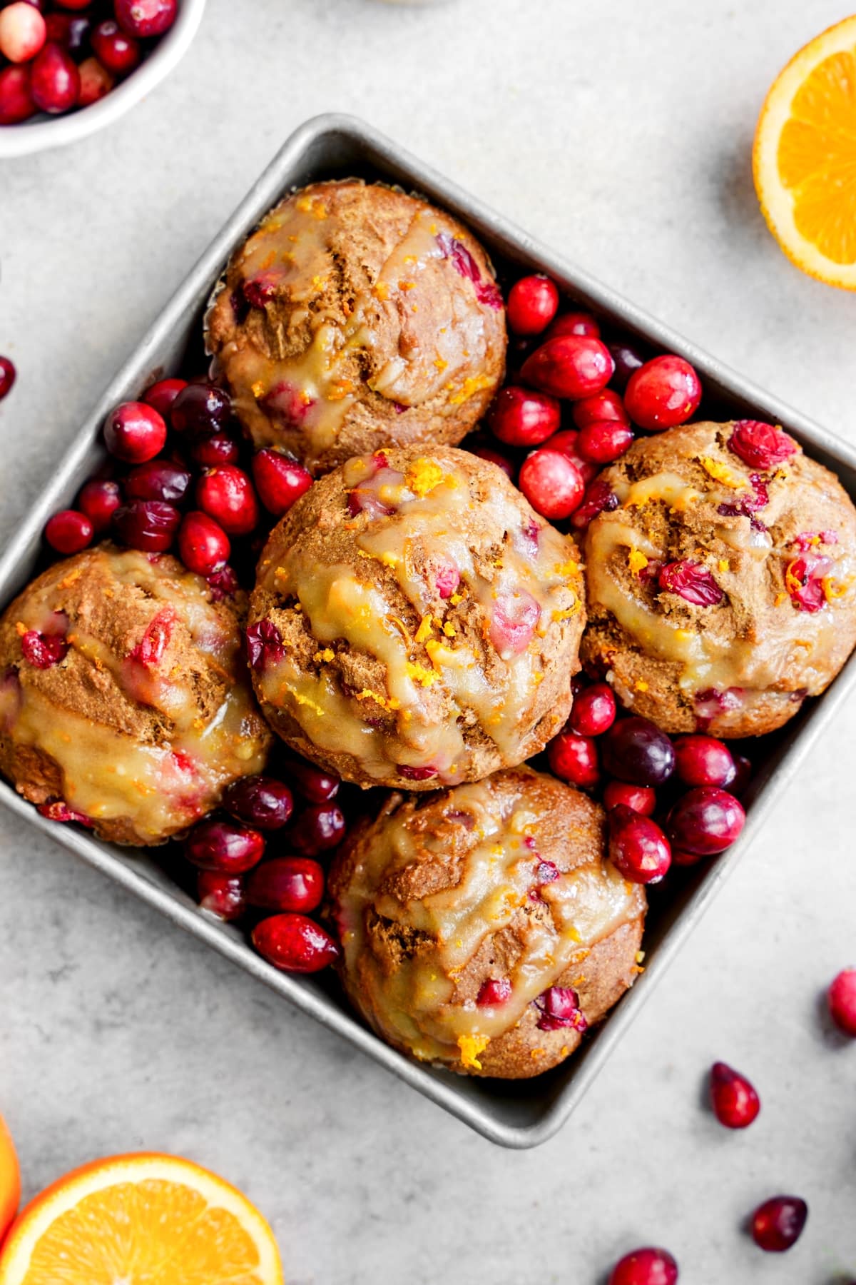 Vegan cranberry orange muffins arranged in a square container, surrounded by fresh cranberries and sliced oranges on a rustic surface