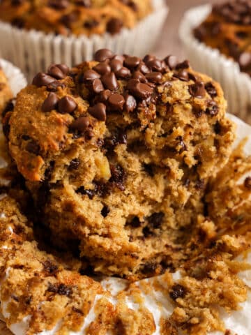 Close-up of a banana chocolate chip protein muffin broken open to reveal its soft, moist, fluffy interior packed with melted chocolate chips. The bakery-style muffin is topped with extra dairy-free chocolate chips and surrounded by crumbs, showcasing its tender texture. Made with ripe bananas and plant-based protein powder, these healthy vegan protein muffins are perfect for breakfast, meal prep, post-workout snacks, or a high-protein treat.