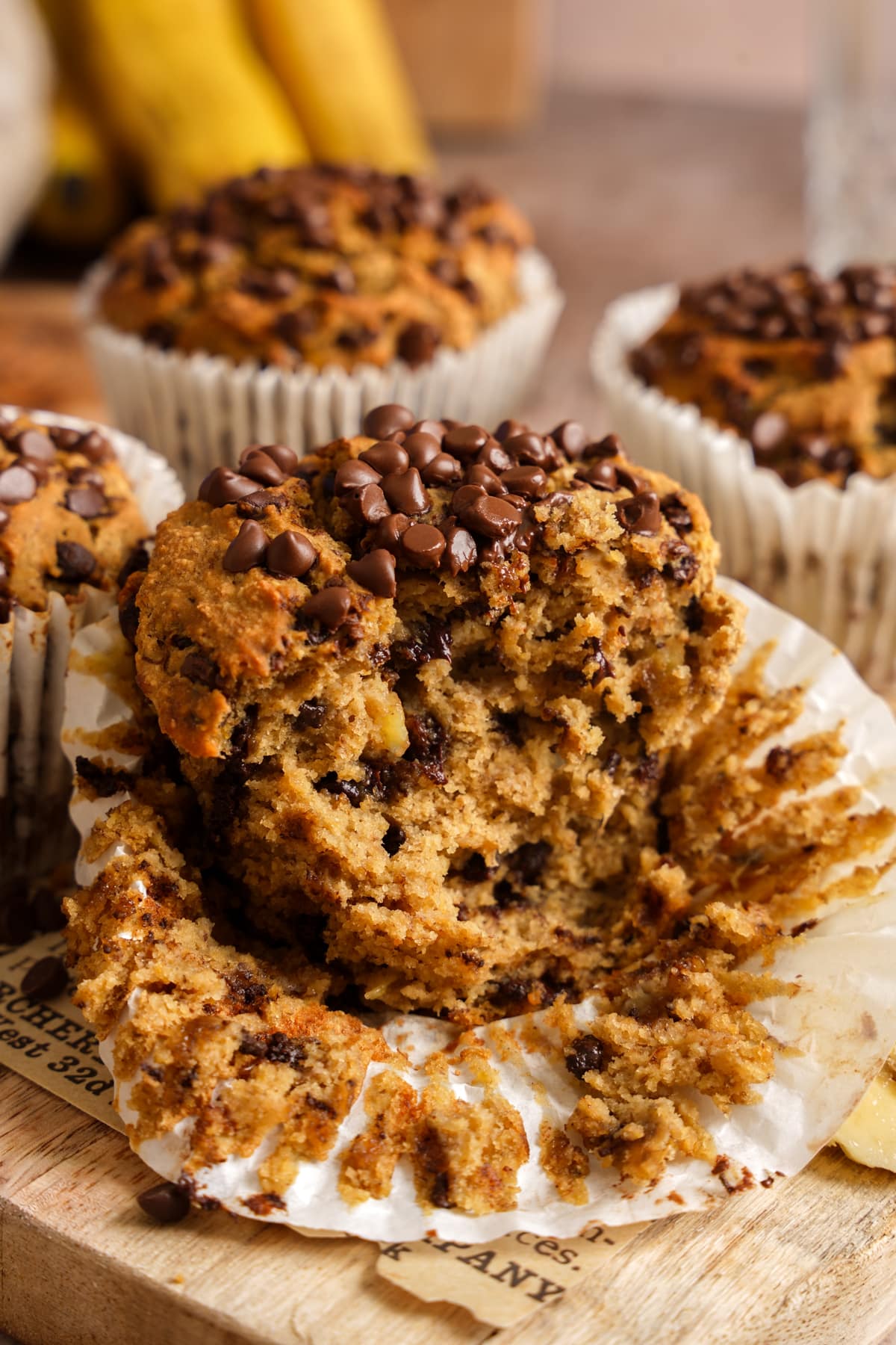 Close-up of a soft and fluffy banana chocolate chip protein muffin broken open to reveal a moist, tender crumb packed with melty chocolate chips. The muffin is topped with extra chocolate chips and surrounded by additional freshly baked muffins in paper liners. Ripe bananas in the background highlight the wholesome ingredients, making this high-protein vegan muffin perfect for breakfast, meal prep, snacks, or a healthy dessert.