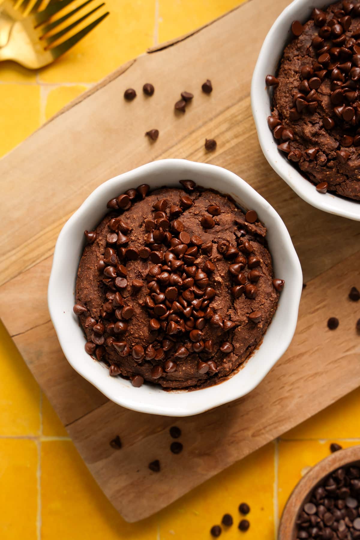 High-protein chocolate chip baked oats served in a white ramekin, topped generously with mini chocolate chips, displayed on a wooden cutting board with scattered chocolate chips and golden forks on a yellow tiled background