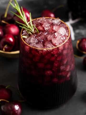 Close-up of a ruby red Sleepy Girl Mocktail filled with crushed ice and garnished with fresh rosemary, surrounded by fresh cherries.