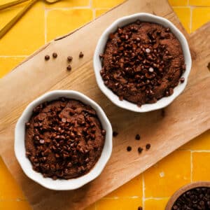 High-protein vegan chocolate baked oats topped with dairy-free chocolate chips, served in white ramekins on a wooden board over a vibrant yellow tiled background. A rich and healthy breakfast or dessert made with Sunwarrior chocolate protein.