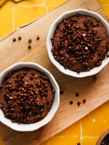 High-protein vegan chocolate baked oats topped with dairy-free chocolate chips, served in white ramekins on a wooden board over a vibrant yellow tiled background. A rich and healthy breakfast or dessert made with Sunwarrior chocolate protein.