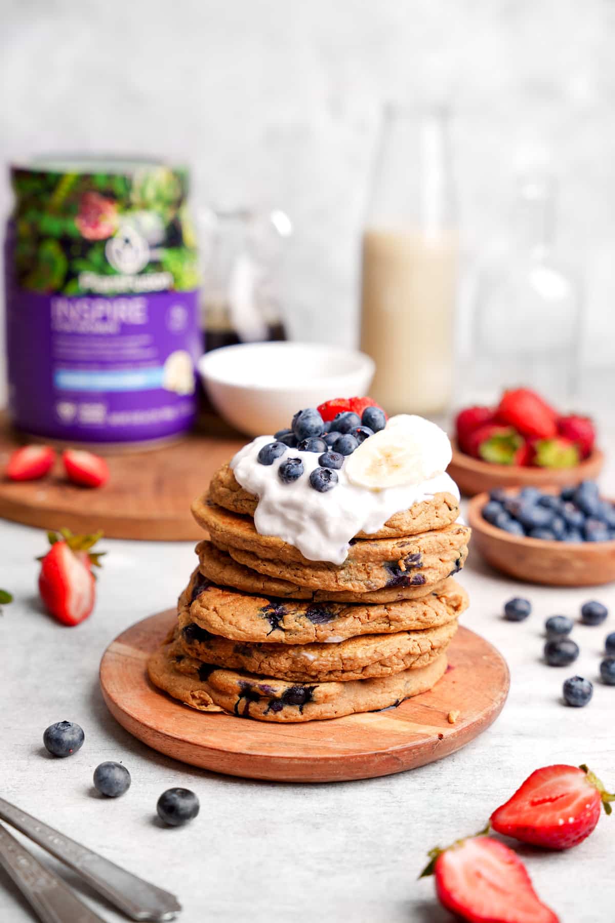 the stack of blueberry protein pancakes with vegan yogurt and fresh fruit on top next to the planttfusion vegan protein powder.