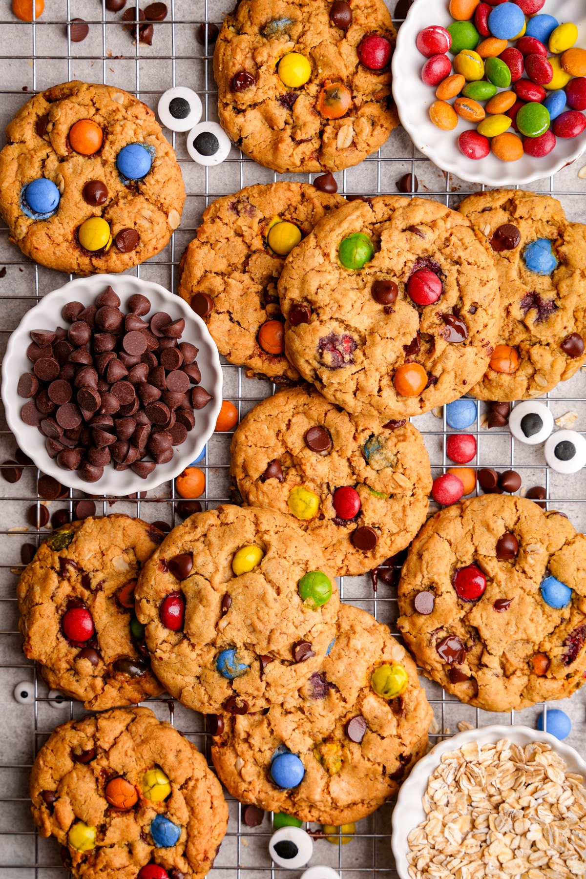 Freshly baked vegan monster cookies loaded with colorful candy-coated chocolates and chocolate chips, cooling on a wire rack beside small bowls of oats, chocolate chips, and vegan candies
