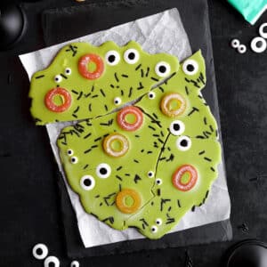 Bright green vegan Halloween candy bark topped with candy eyeballs, orange gummy rings, and black sprinkles, broken into fun pieces.