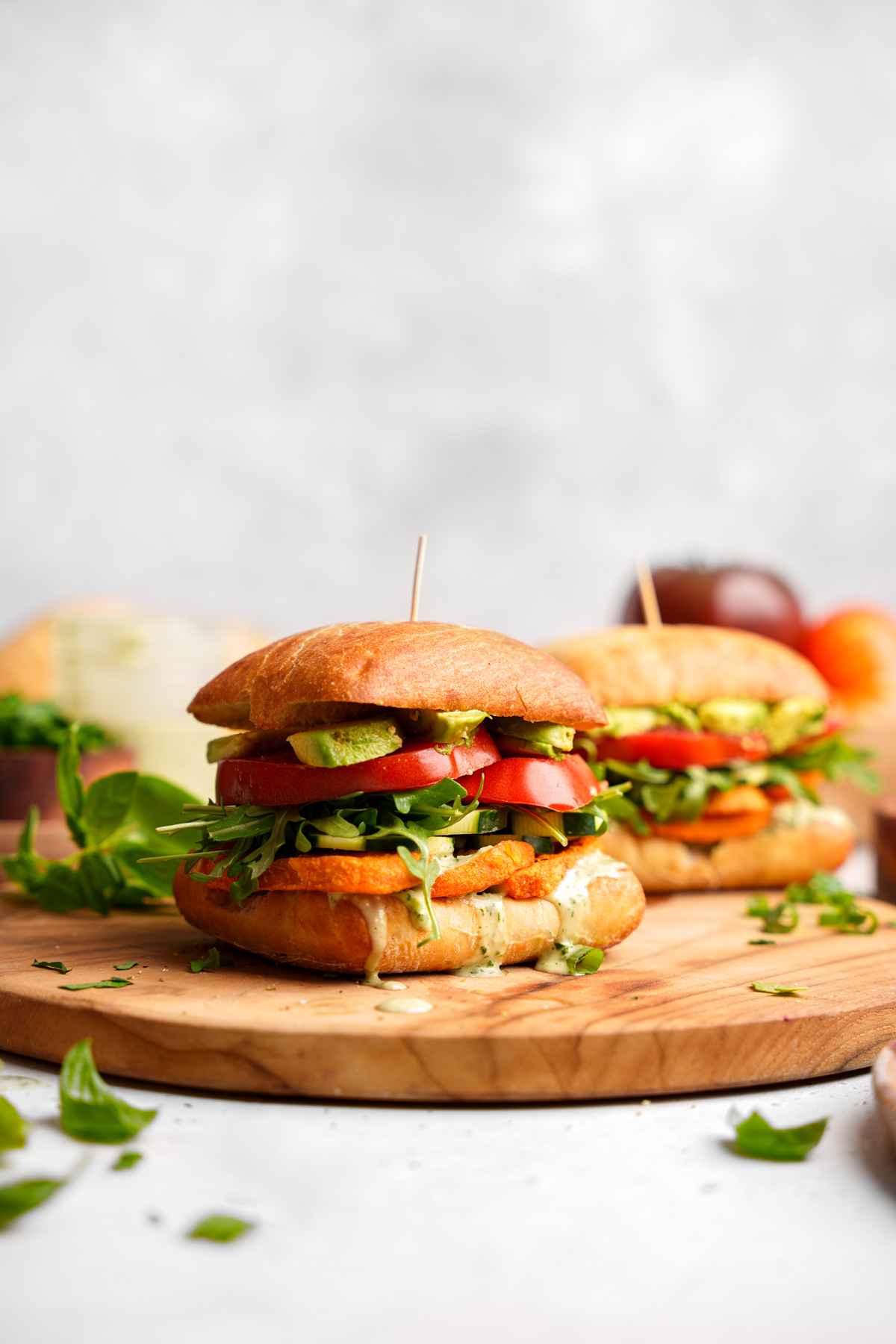 the vegan sweet potato sandwiches on a wooden cutting board with fresh herbs around them and the green goddess dressing oozing out of them