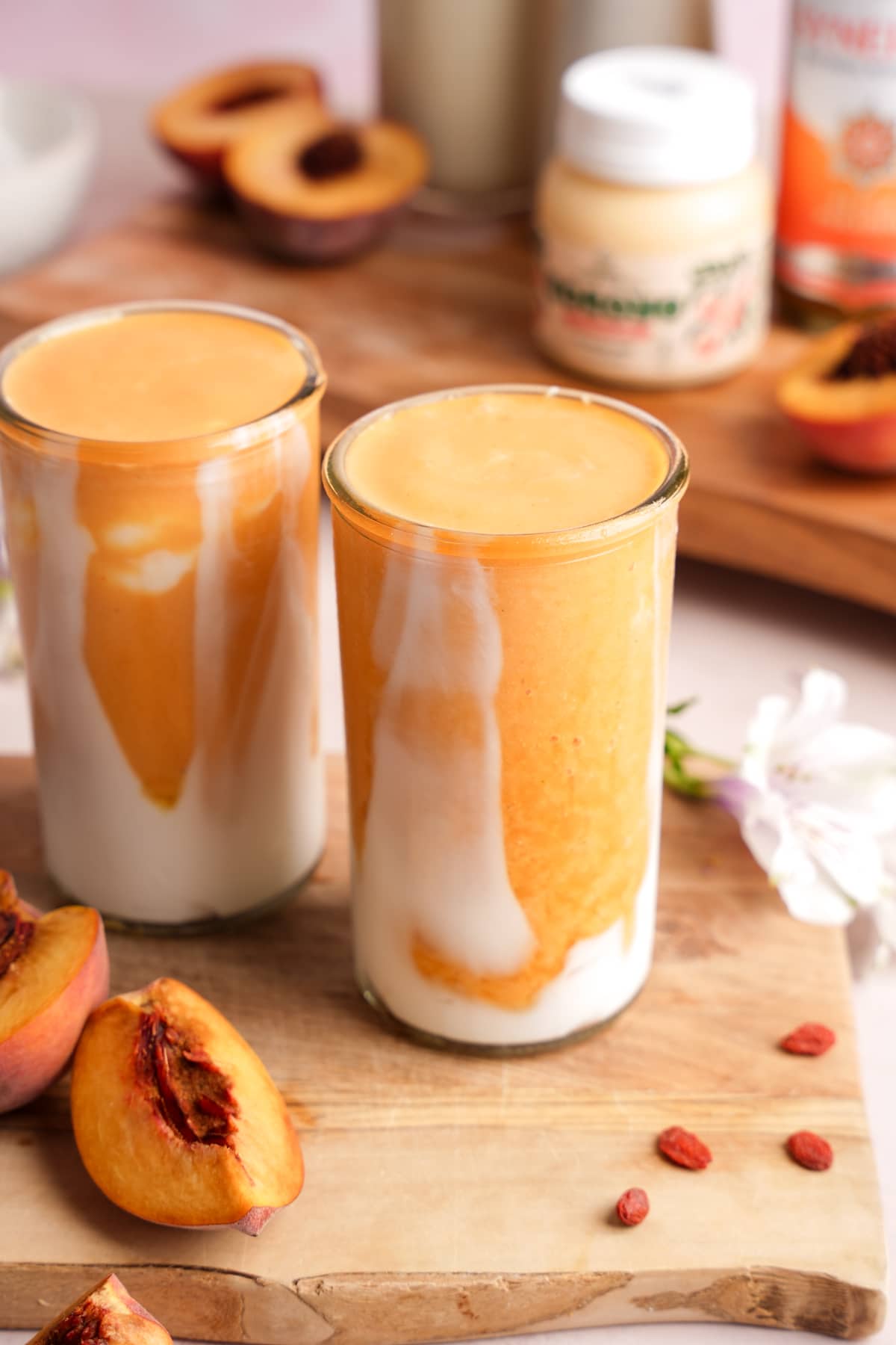 Peach smoothie layered with creamy coconut yogurt in tall clear glasses, surrounded by fresh peach slices, goji berries, and superfood ingredients on a wooden board.