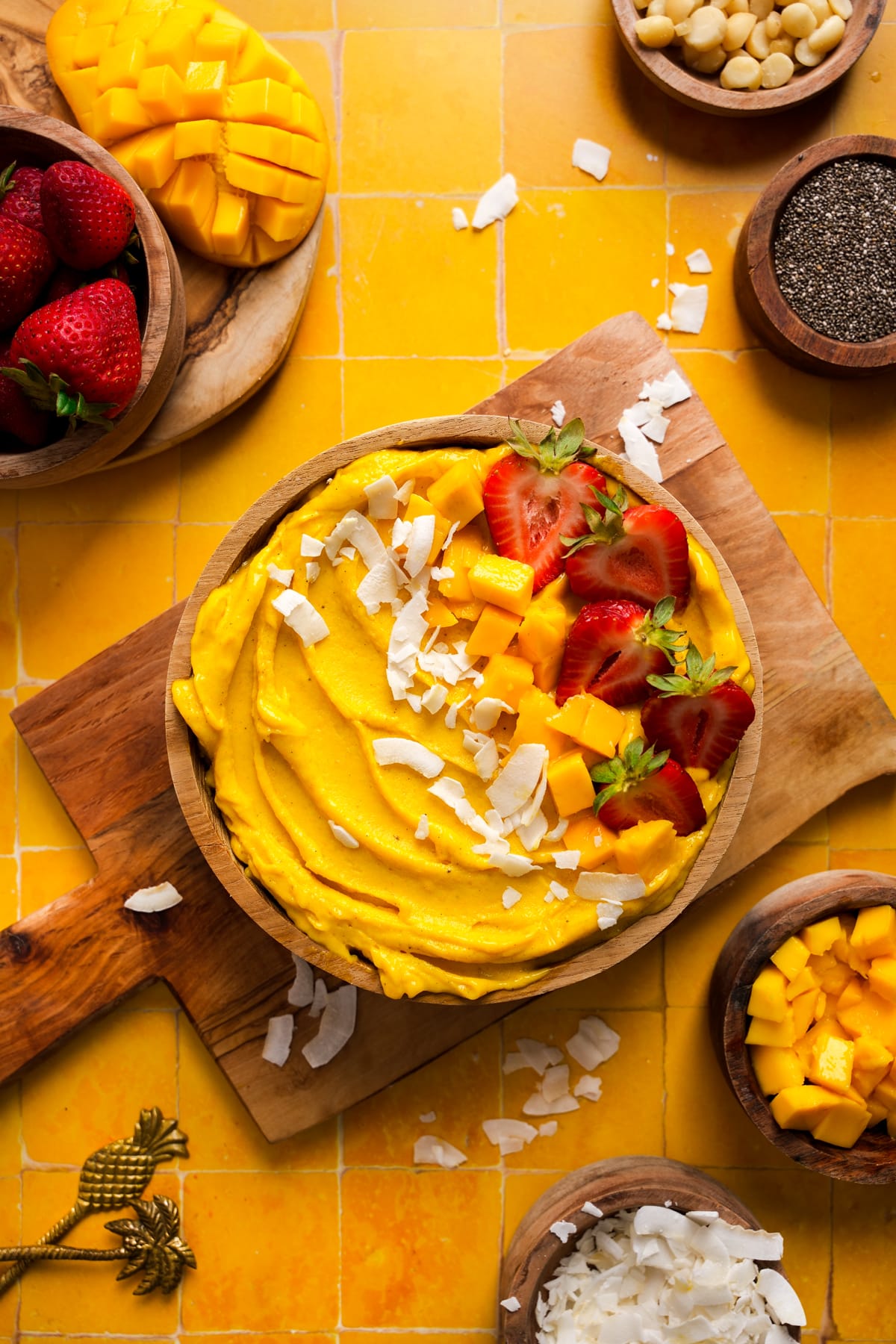 Tropical mango smoothie bowl topped with diced mango, sliced strawberries, and coconut flakes, served in a wooden bowl on a cutting board over a vibrant yellow tile background, surrounded by bowls of fresh fruit, macadamia nuts, chia seeds, and coconut.