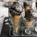 Close-up of two waffle cones filled with rich, dark black sesame ice cream, topped with black sesame seeds, displayed upright in clear glasses within a rustic wire rack.