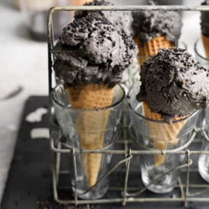 Close-up of two waffle cones filled with rich, dark black sesame ice cream, topped with black sesame seeds, displayed upright in clear glasses within a rustic wire rack.