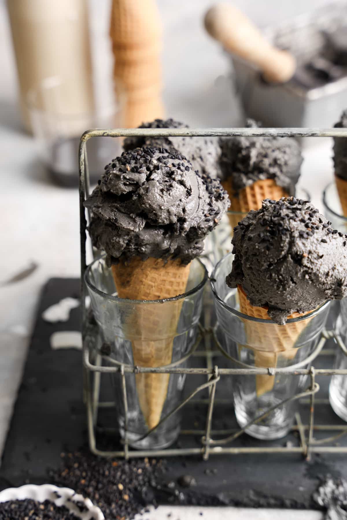 Creamy scoops of black sesame ice cream served in waffle cones, placed upright in clear glasses within a rustic wire holder, with black sesame seeds scattered on the surface below.