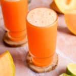 Vibrant glass of cold-pressed cantaloupe juice topped with natural foam, sitting on a wooden coaster, surrounded by fresh cantaloupe wedges and lime halves on a soft pink surface.