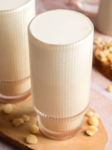 Glass of creamy homemade macadamia nut milk on a wooden serving board, surrounded by chopped raw macadamia nuts with a soft pink backdrop.